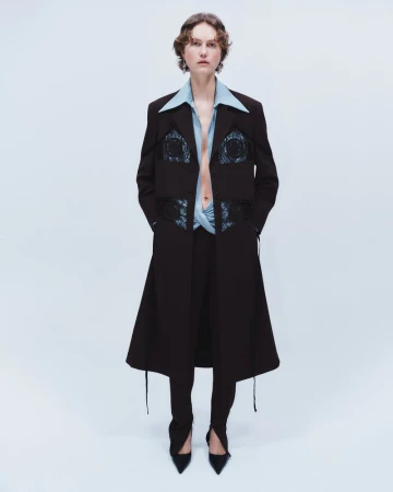 Julie Hoomans wearing a black tailored long coat and light blue collared shirt for Beaufille Pre Fall 2024 against a pale backdrop.