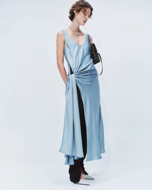 Fashion model poses in a studio wearing a blue satin dress with a knotted detail, paired with black trousers and silver high heels.