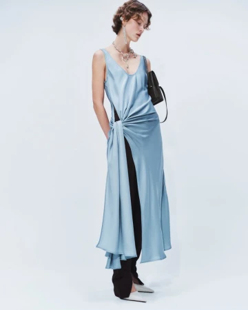 Fashion model poses in a studio wearing a blue satin dress with a knotted detail, paired with black trousers and silver high heels.