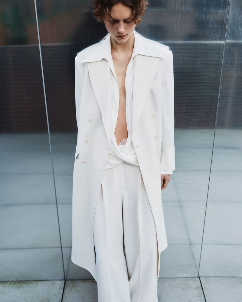 A young adult woman stands in a neutral outfit, featuring a white coat and wide-legged pants, against a grey mirrored urban background.