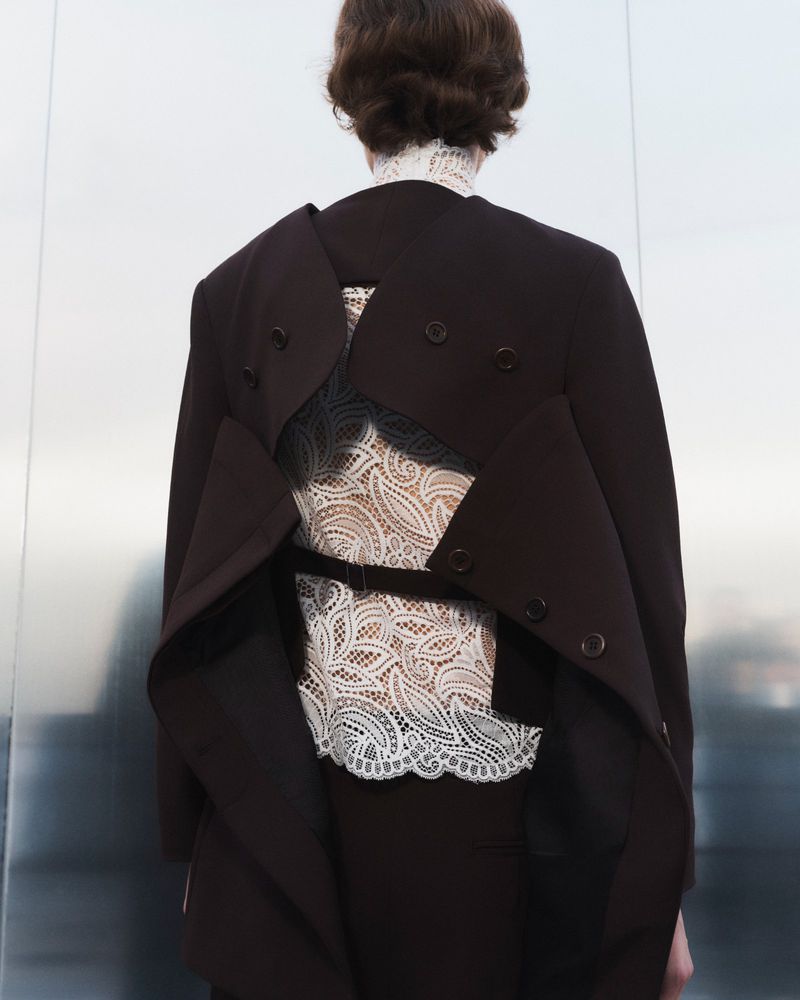 Back view of person wearing brown coat with lace detail, against a silver backdrop. Fashion shot with focus on clothing texture and design.