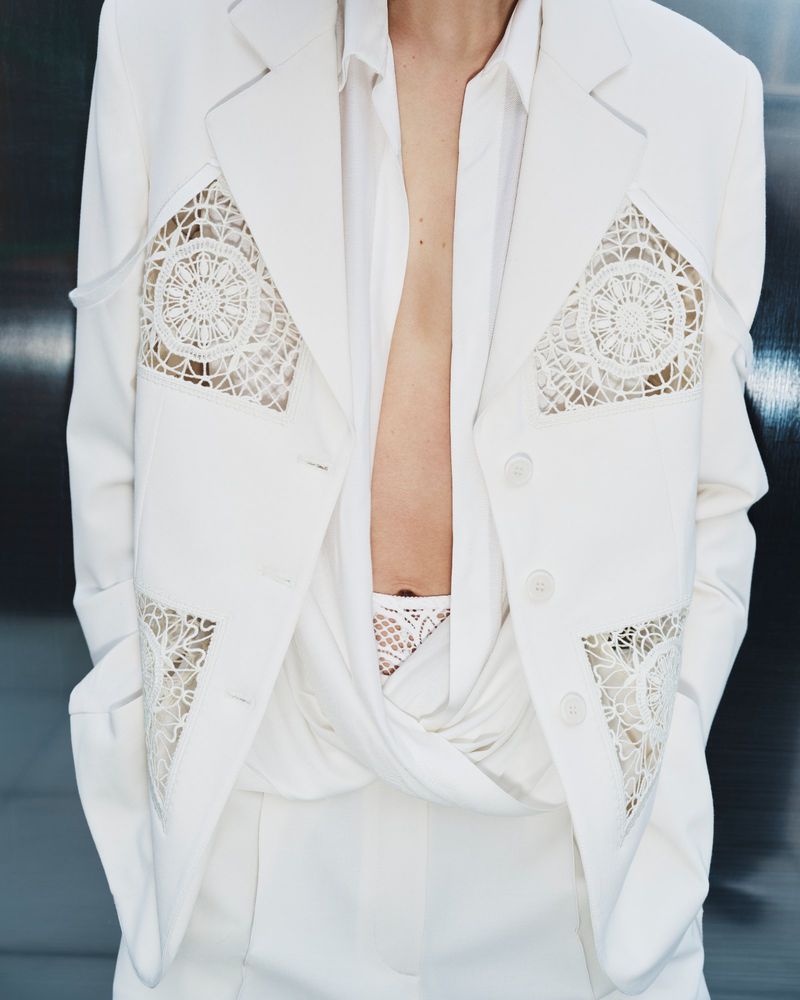 Person in a white suit with lace cutouts posing against a dark background. The tailored jacket is buttoned and worn with a white shirt.
