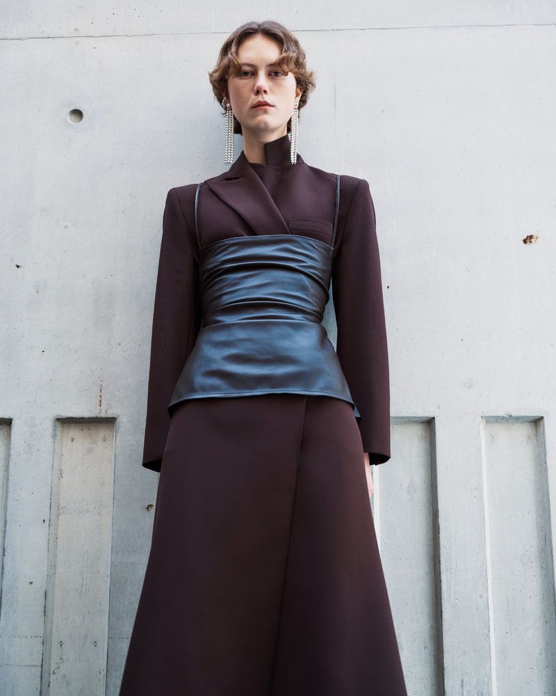 Fashion shot of woman with wavy hair wearing a brown skirt and blazer with a black leather corset in front of a concrete wall.