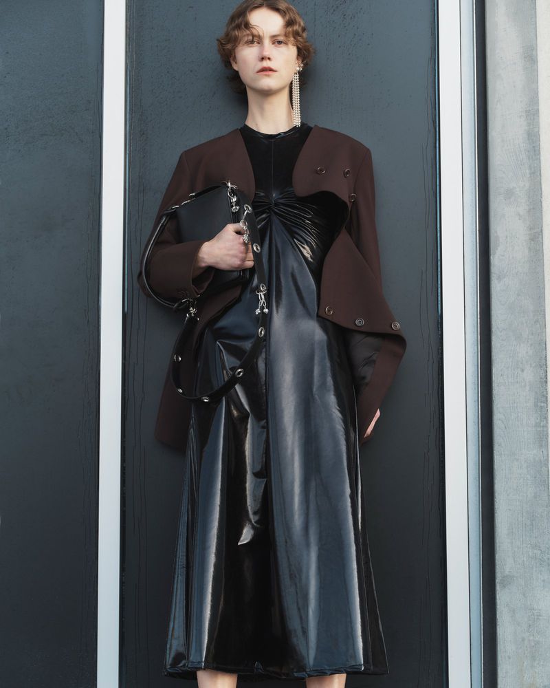 Fashionable woman poses in a black dress and brown jacket against a dark grey paneled wall, accessorized with earrings and a handbag.