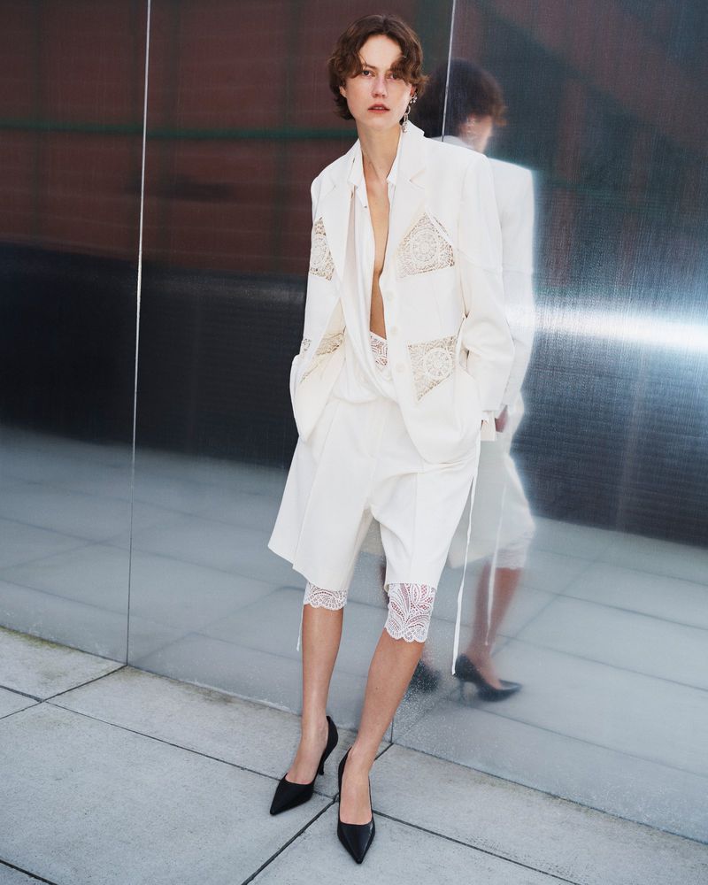 Model in white blazer and matching shorts with lace detailing standing against a reflective glass surface.