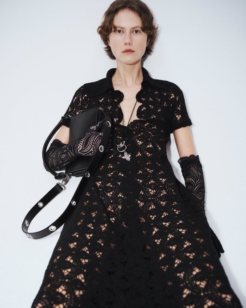 A woman in a black lace dress poses against a white background, accessorized with a matching black handbag and lace gloves for fashion.