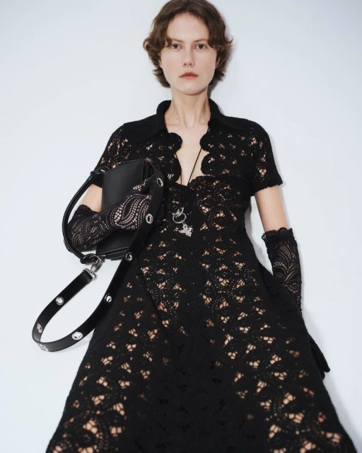 A woman in a black lace dress poses against a white background, accessorized with a matching black handbag and lace gloves for fashion.