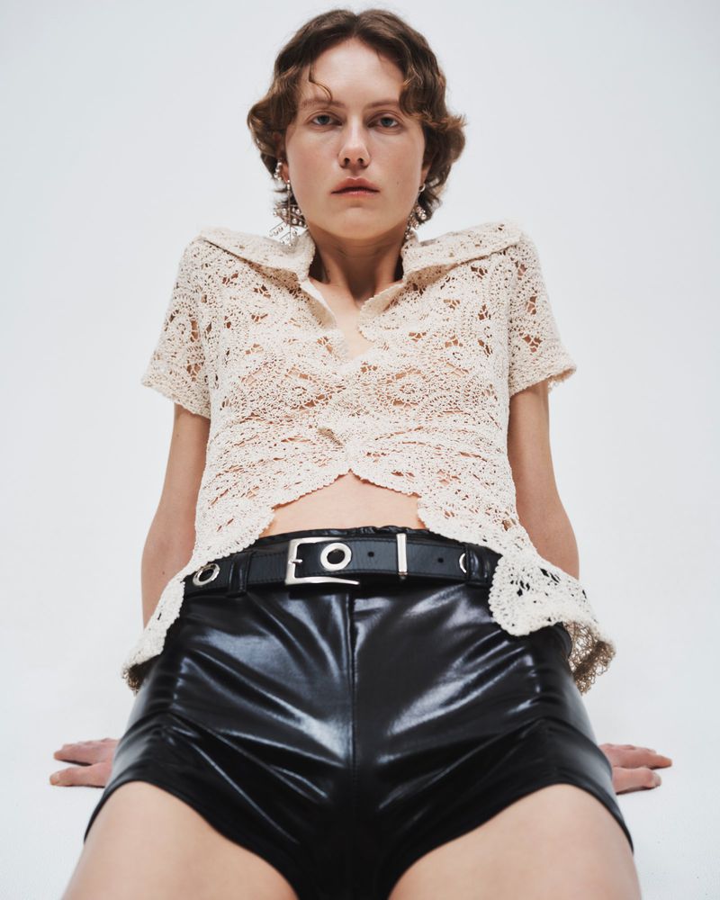 A young woman poses in a cream crochet top and shiny black shorts in a studio setting, under bright, even lighting.