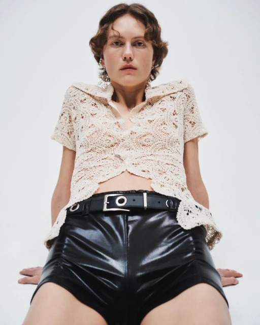 A young woman poses in a cream crochet top and shiny black shorts in a studio setting, under bright, even lighting.