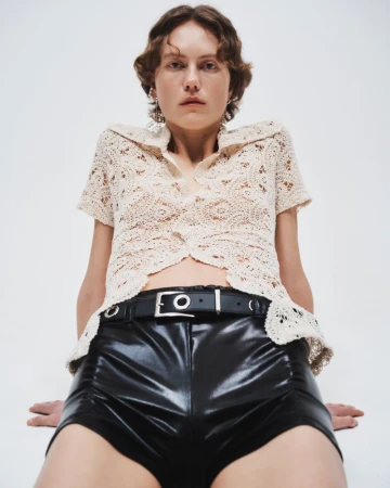 A young woman poses in a cream crochet top and shiny black shorts in a studio setting, under bright, even lighting.