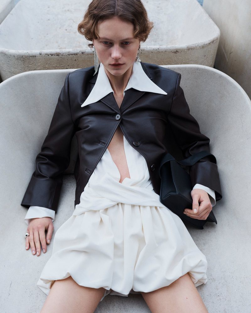 Fashion editorial shot of a woman in a black jacket and white dress, posing in a concrete basin with natural light and soft color grading.