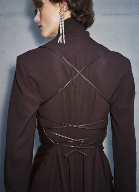 Back view of a slender adult woman wearing a dark brown dress with long sleeves and a criss-cross leather strap design.