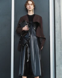 Model in a shiny black dress and brown blazer holding a black handbag against a dark background