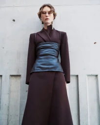 Model wearing a dark structured blazer and leather corset top against a raw concrete architectural wall