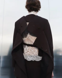Model seen from behind wearing a deconstructed dark open-back blazer over a white floral lace top against reflective metal panels.