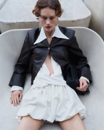 Model Julie Hoomans reclining in a dark leather jacket and draped white mini dress inside a sculpted concrete basin.