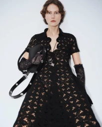 Model wearing a black lace dress and matching gloves, holding a leather handbag against a plain white background.