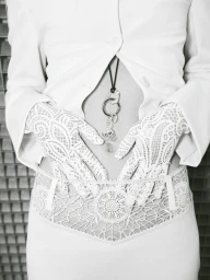 Black and white close-up of a model wearing intricate white lace gloves, an unbuttoned jacket, and a key ring pendant necklace.