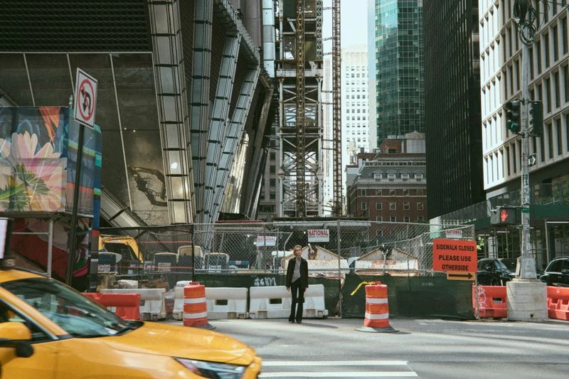 Urban street scene with a pedestrian in a dark coat standing near a construction site, taxi in foreground, and tall buildings in the background.
