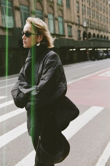 Woman in sunglasses and puffy jacket crossing a city street. Urban setting with buildings in the background, captured in medium shot.