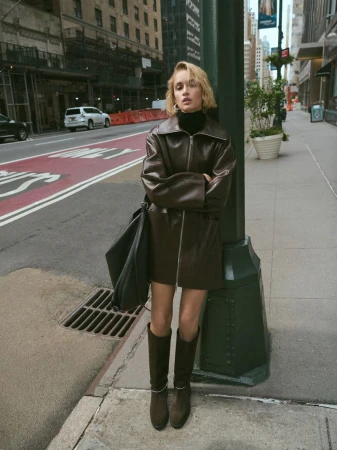 A fair-skinned young woman poses on a city sidewalk in a brown leather jacket and knee-high boots with buildings in the background.
