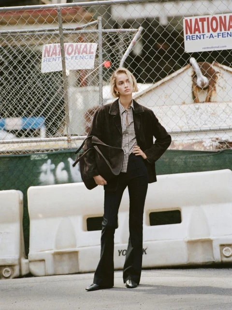 A stylish adult woman stands outdoors in a brown jacket, checkered shirt, and dark pants, carrying a duffel bag on her shoulder.