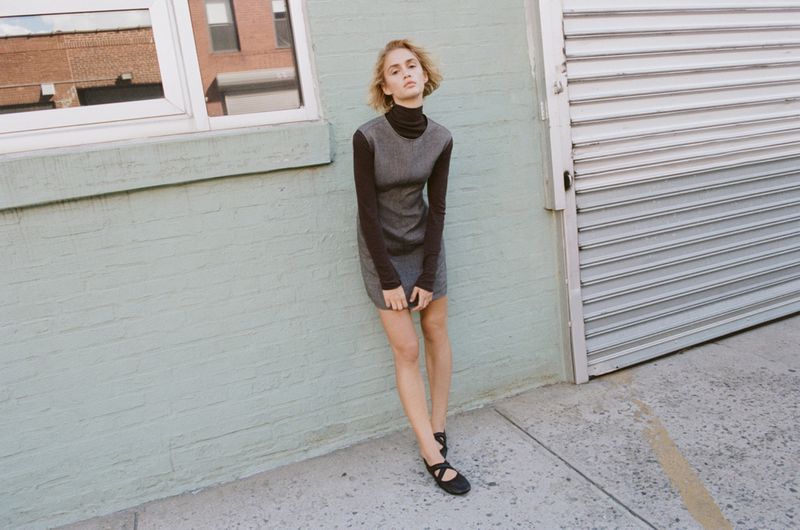 Young woman in long-sleeve top and gray dress standing in front of a pale green brick wall with an aluminum roll-up door.
