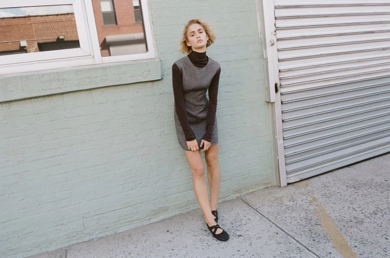 Young woman in long-sleeve top and gray dress standing in front of a pale green brick wall with an aluminum roll-up door.