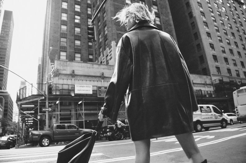 Woman in a black leather coat walks in a city street with tall buildings, holding a black umbrella; monochromatic, low-angle shot.