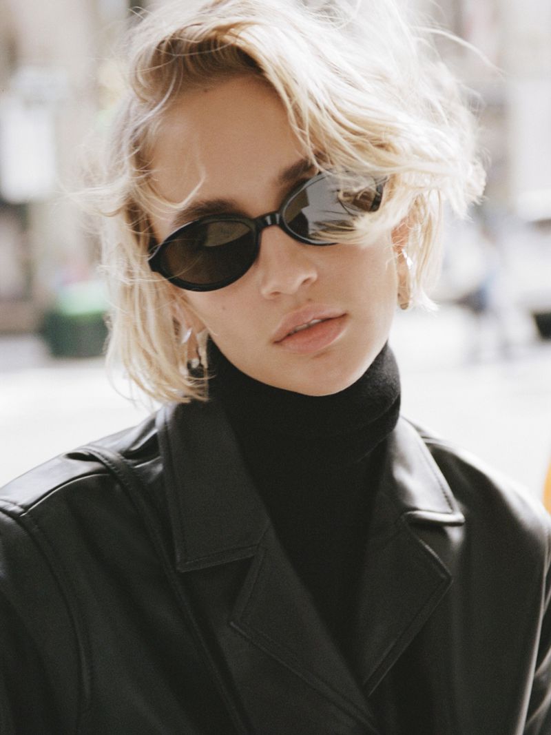 Model wearing dark oval sunglasses and a black leather jacket with a turtleneck against an urban street background.