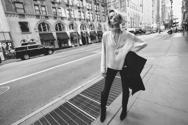 Black and white fashion shot of a young adult woman posing on a city street in a blouse, dark pants, and boots, with buildings behind.