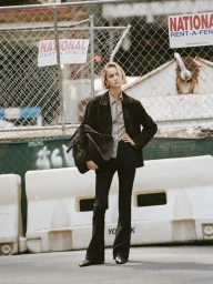 Model Quinn Mora wearing a dark jacket and trousers holding a large handbag in front of a construction fence