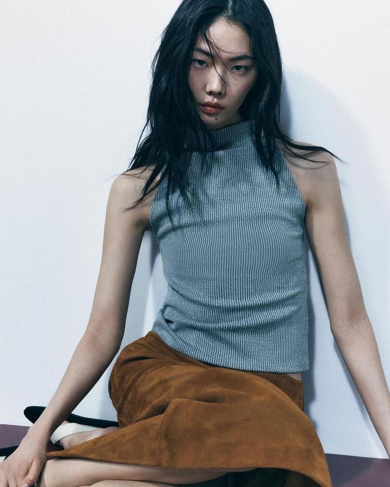 A woman with dark hair poses indoors, wearing a grey ribbed top and a brown suede skirt, against a plain white wall backdrop.