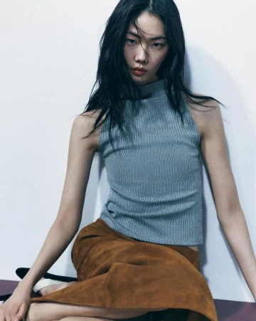 A woman with dark hair poses indoors, wearing a grey ribbed top and a brown suede skirt, against a plain white wall backdrop.