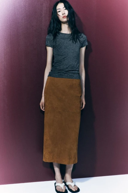 A full-length studio shot of an Asian female fashion model in a shiny top and suede skirt, posing against a muted maroon background.