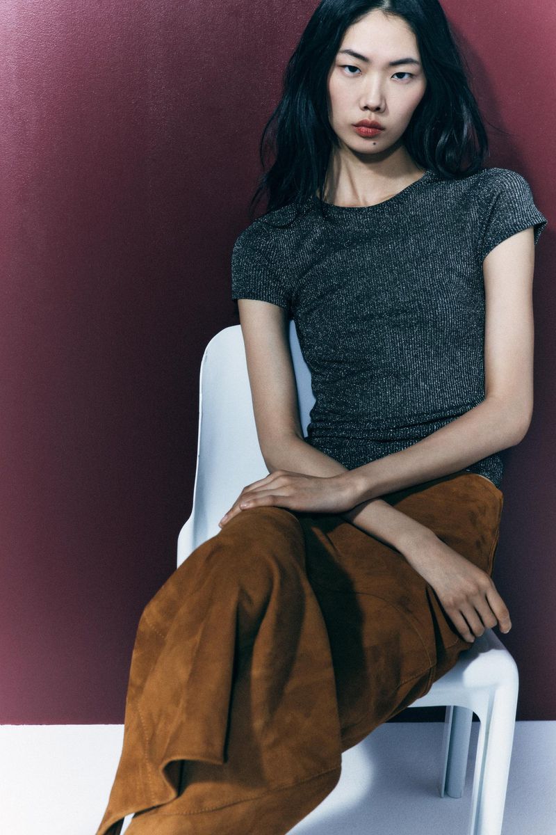 Elegant Asian woman in a dark grey shirt and brown suede skirt sitting on a white chair against a dark reddish background, fashion shot.