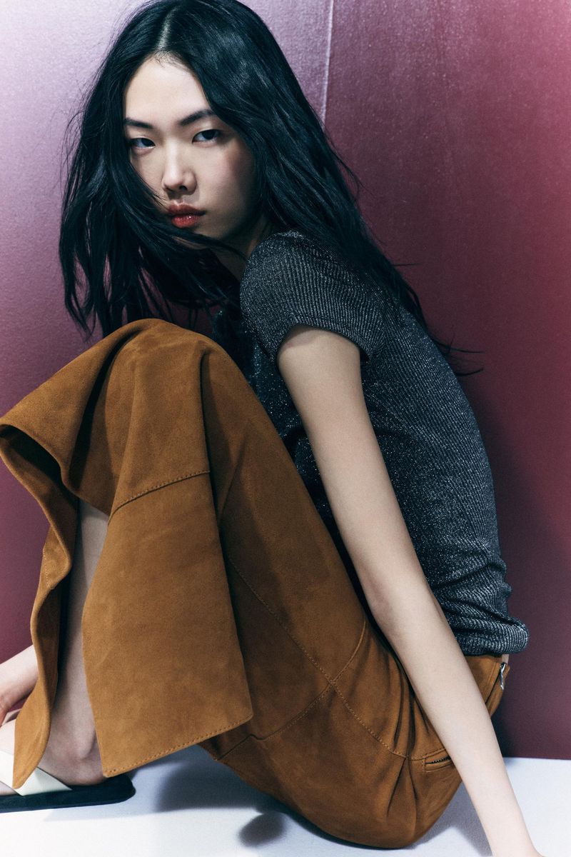 Indoor studio shot features an Asian woman in a grey top and brown pants, seated against a maroon backdrop. Fashion editorial portrait.