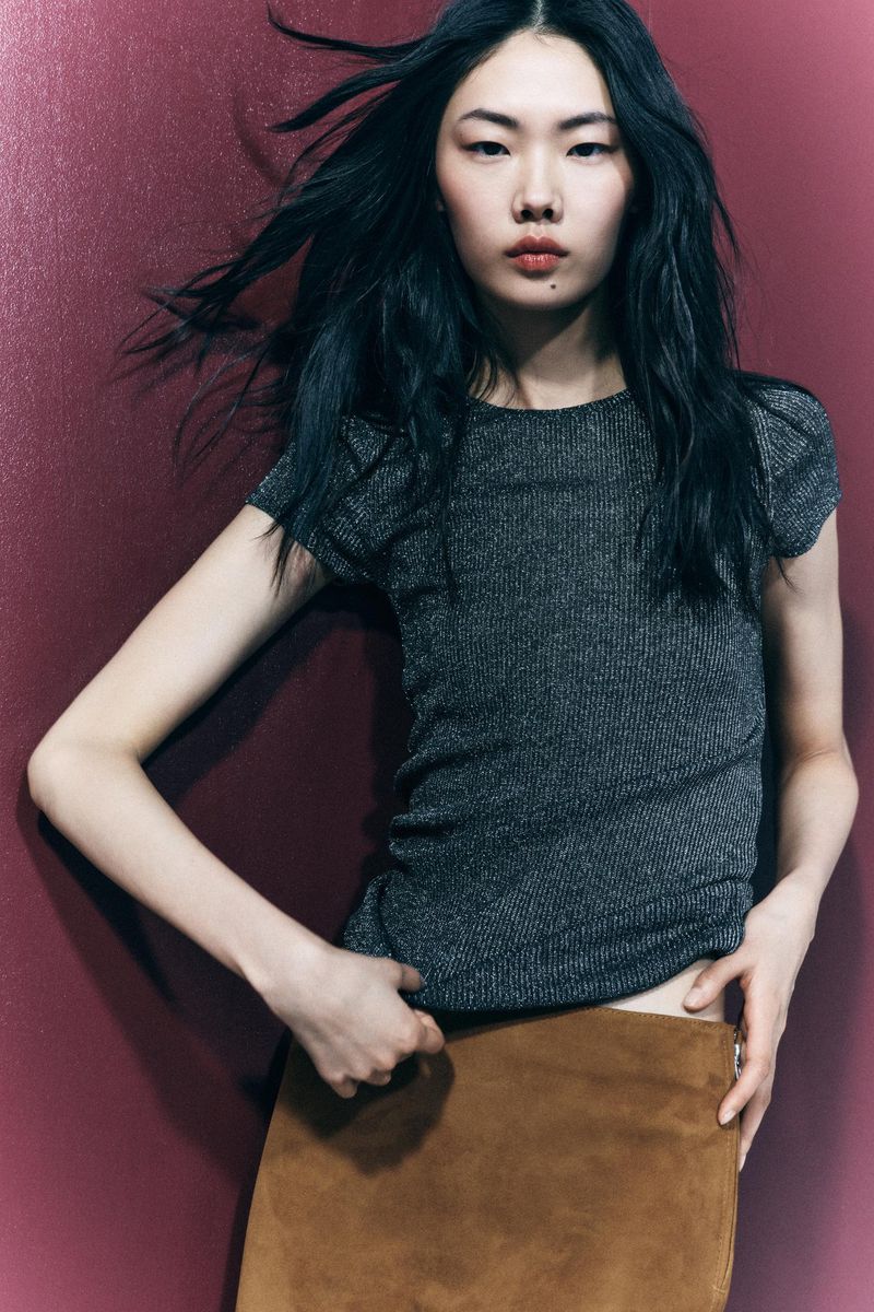 Fashion shot of an Asian woman with long black hair in a gray ribbed top and brown skirt against a maroon background in portrait orientation.