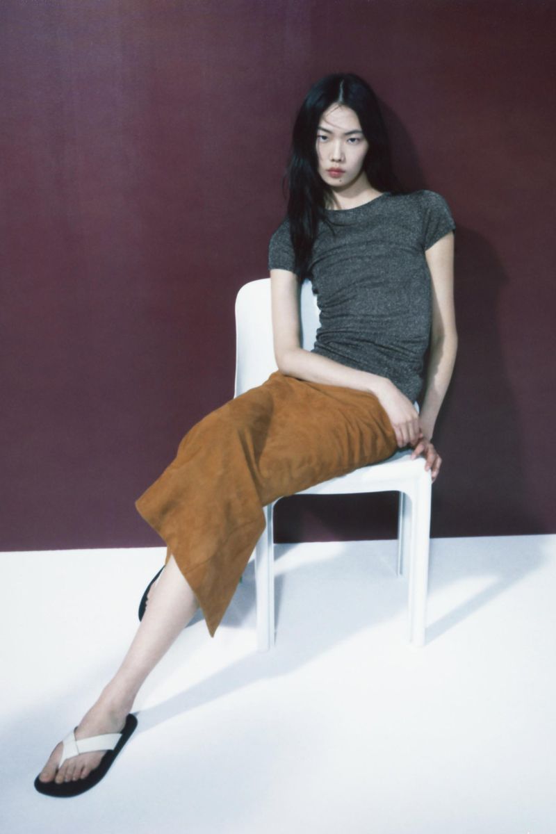A stylish Asian woman sits on a white chair against a muted purple backdrop wearing a grey top, brown skirt, and white sandals.