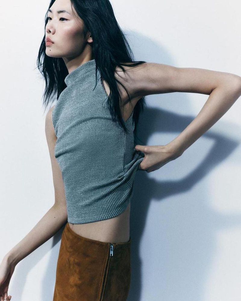 A slender Asian woman in a gray ribbed top and tan suede skirt adjusts the top, posing against a light gray background in a studio setting.