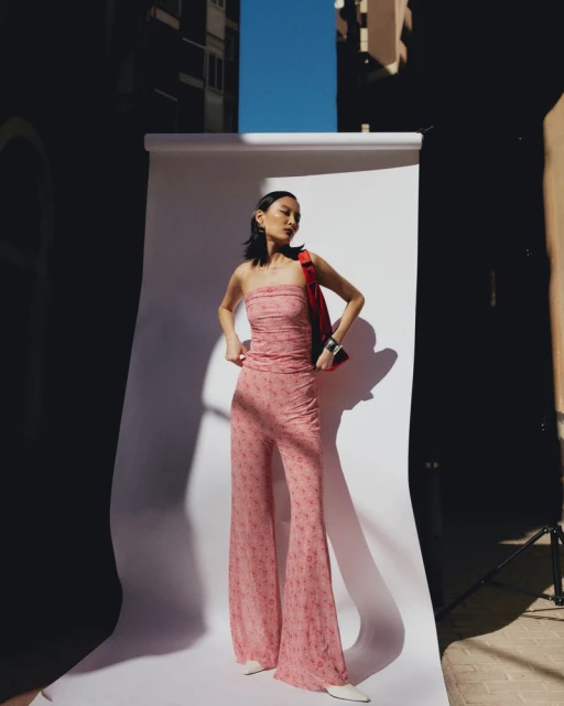 An Asian woman poses in a pink floral two-piece outfit against a white backdrop, with surrounding buildings and bright blue sky visible.