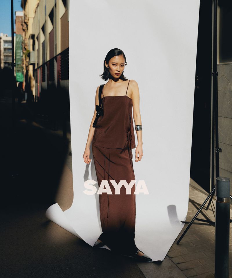 Asian model in a brown spaghetti-strap dress posing in front of a backdrop next to a city street with buildings and natural lighting.
