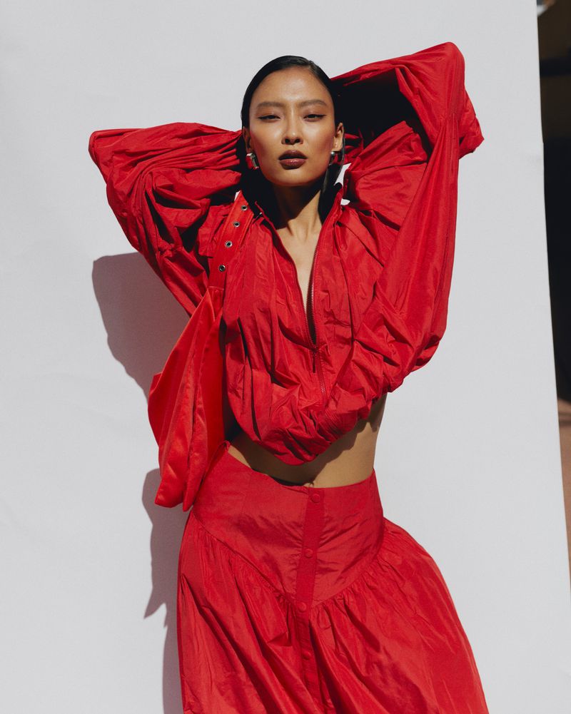 Asian woman in vibrant red outfit strikes a pose against a neutral wall in a studio setting, creating a striking visual contrast.