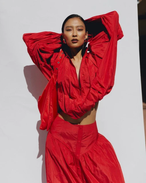 Asian woman in vibrant red outfit strikes a pose against a neutral wall in a studio setting, creating a striking visual contrast.