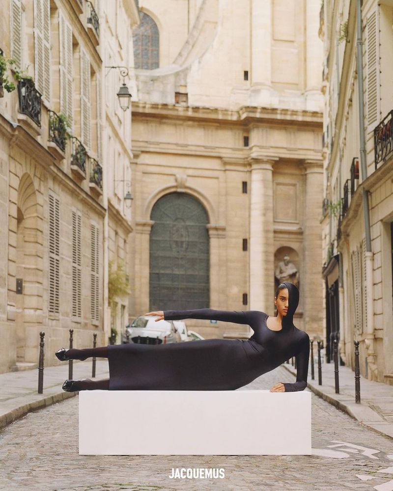 Fashion image on city street featuring a woman in a dark bodysuit reclining on a white block with historic architecture behind her.