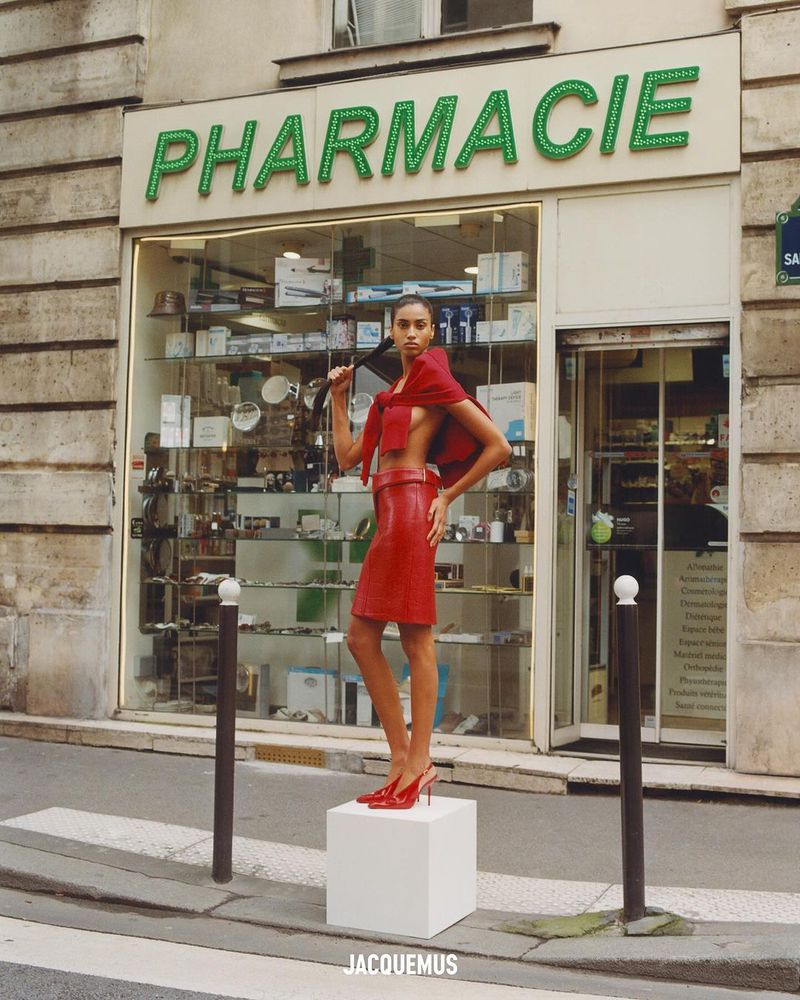 A fashion model poses on a white block outside a Paris pharmacy wearing a red leather skirt and top, striking a stylish pose.