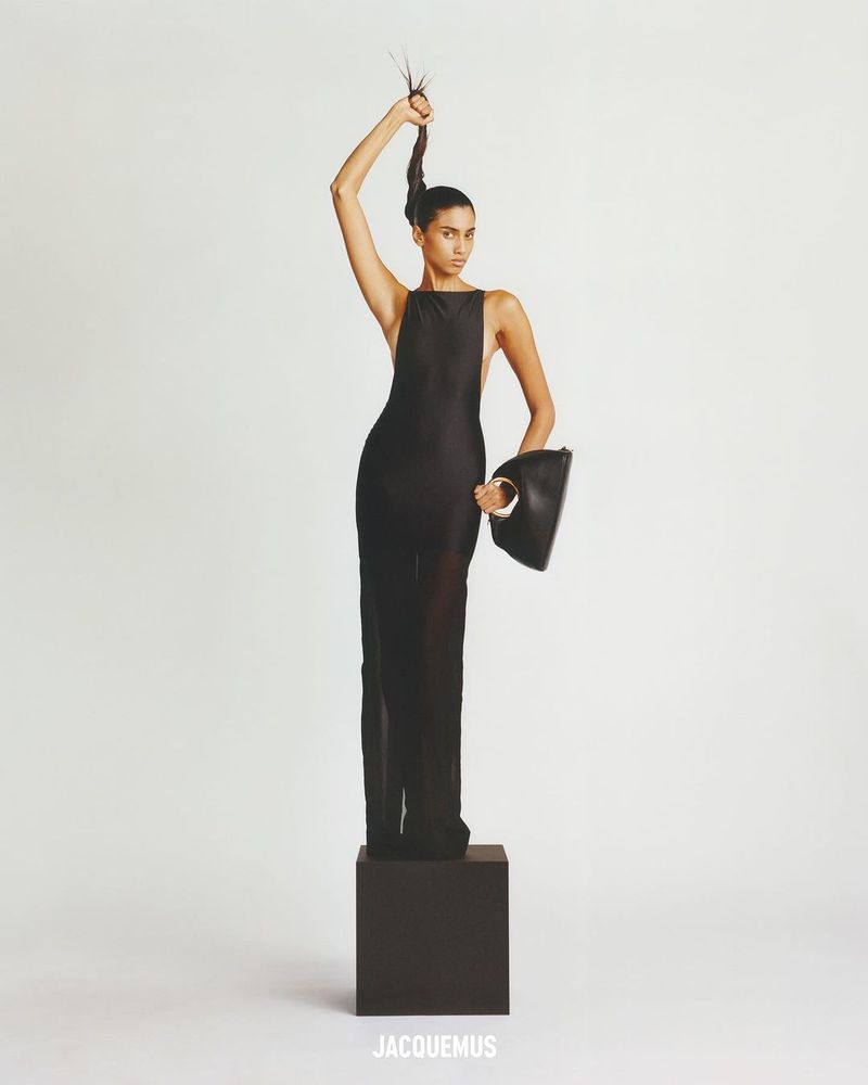 A slender adult woman in a sleek black dress stands atop a dark cube, posing with one arm raised, holding her hair. Fashion shot.