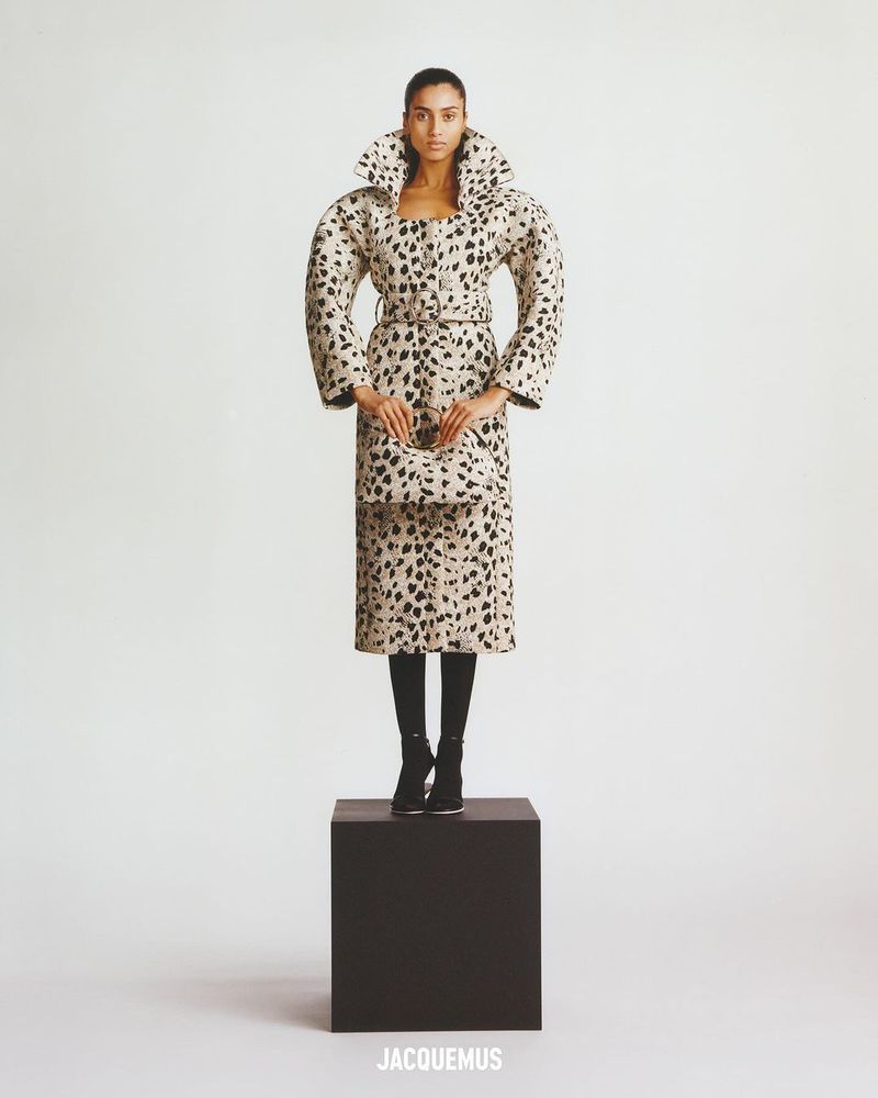 Full-length studio shot of a woman in an oversized, printed coat and skirt with matching handbag, standing on a black cube.