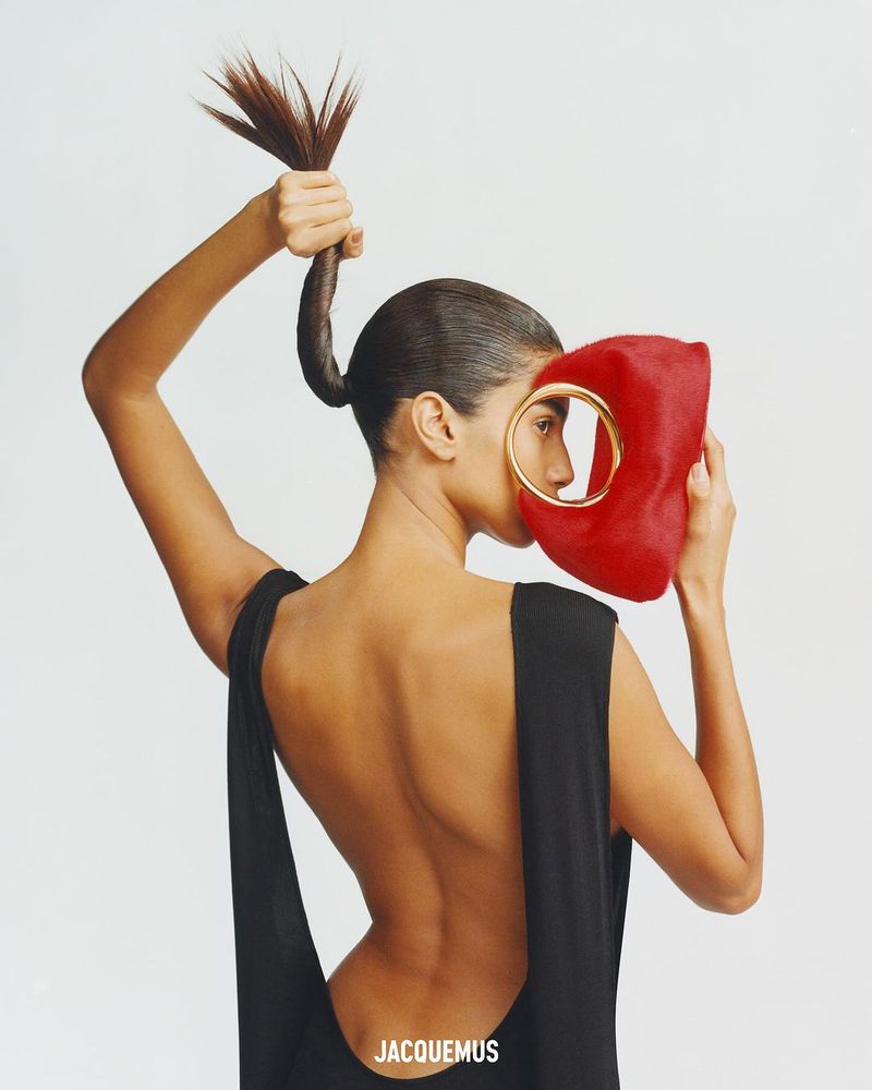 Woman wearing a black backless dress holds up her ponytail and a red handbag with gold handle against a plain, neutral background.