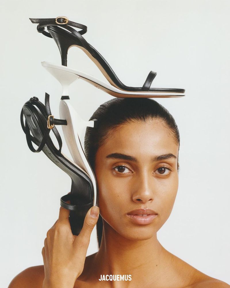 Fashion shot of a young woman in a studio holding black and white stiletto high heels. The shoes are balanced on top of her head.